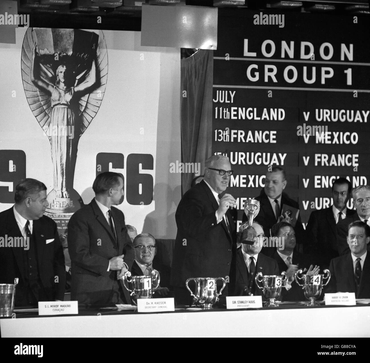 Sir Stanley Rous, President of FIFA, holding the World Championship Jules Rimet Cup at the Royal Garden Hotel in Kensington, where he presided over the draw for the competition to be held in England in July. With him at the table are (l-r) SL Mordy Maduro, Helmet Kaeser, Harry H Cavan, and the Earl of Harewood. Stock Photo