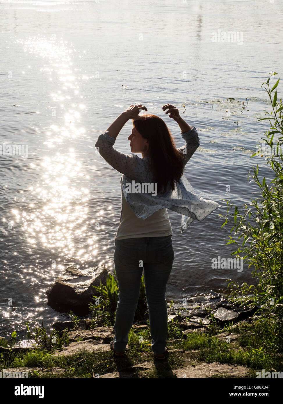Woman standing by river with arms raised and scarf blowing in wind Stock Photo