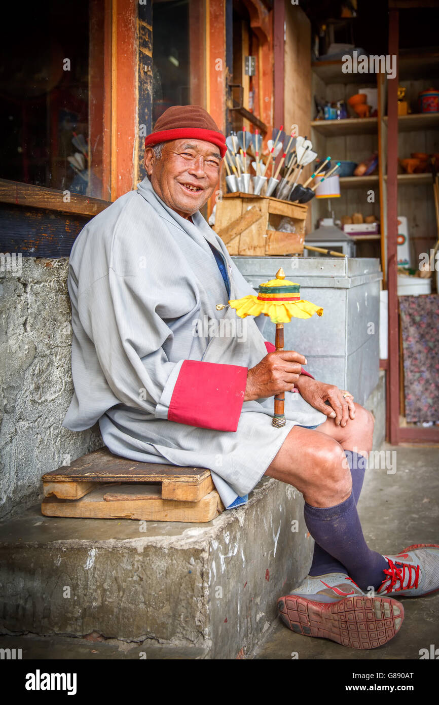 Older Man sitting in front of shop dressed in Bhutanese traditional clothing spins prayer wheel with cloth. Stock Photo