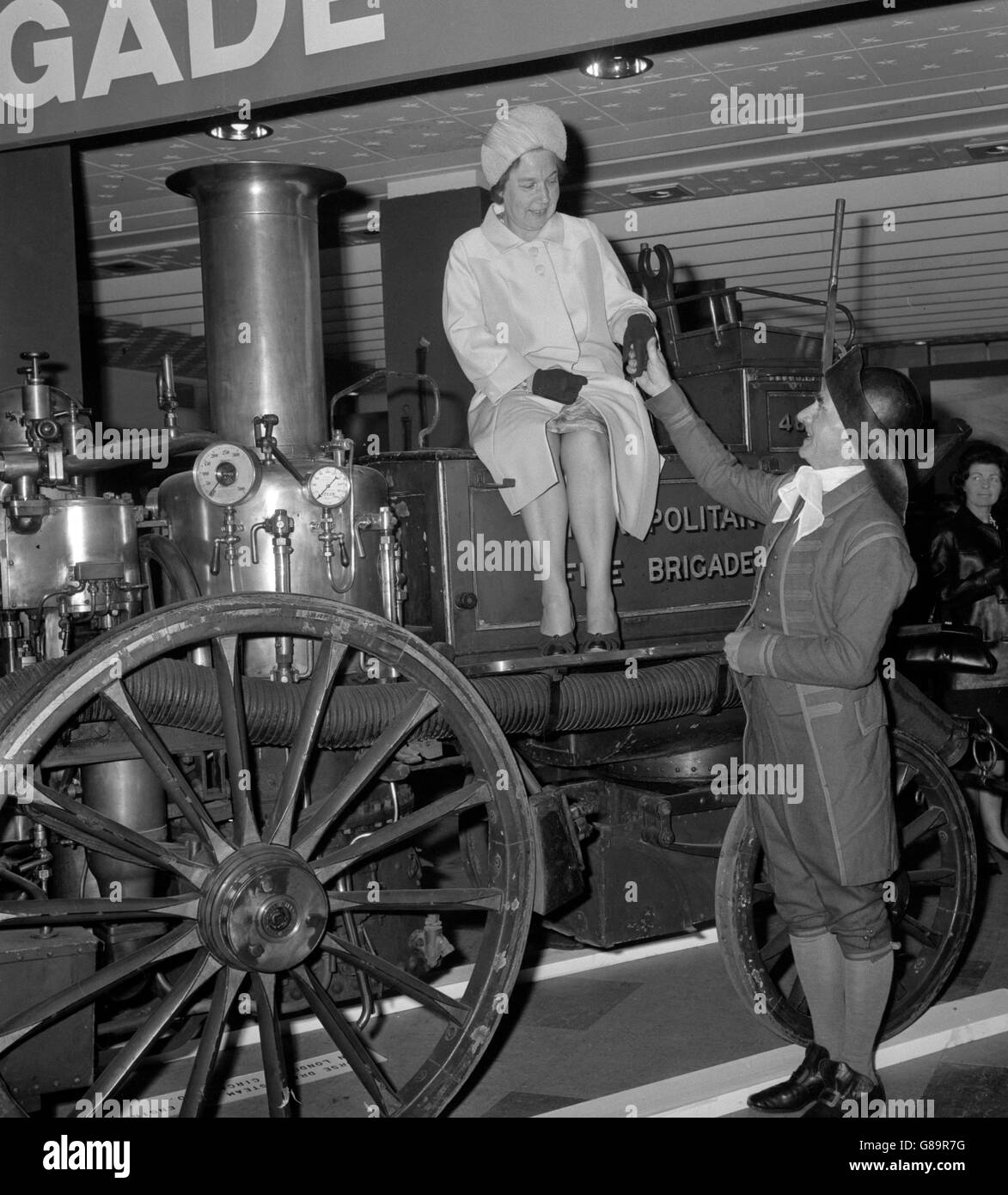 Alice Bacon, CBE, Minister of State, Home Office, tries the seat on an 1860 horse-drawn fire engine when she opened the London Fire Brigade's Centenary Exhibition at Selfridge's in Oxford Street, London. Explaining the exhibit is Crofton Preen, wearing the Phoenix Insurance Company Fire Uniform of the early 18th century, when the insurance companies were responsible for fire-fighting. Stock Photo