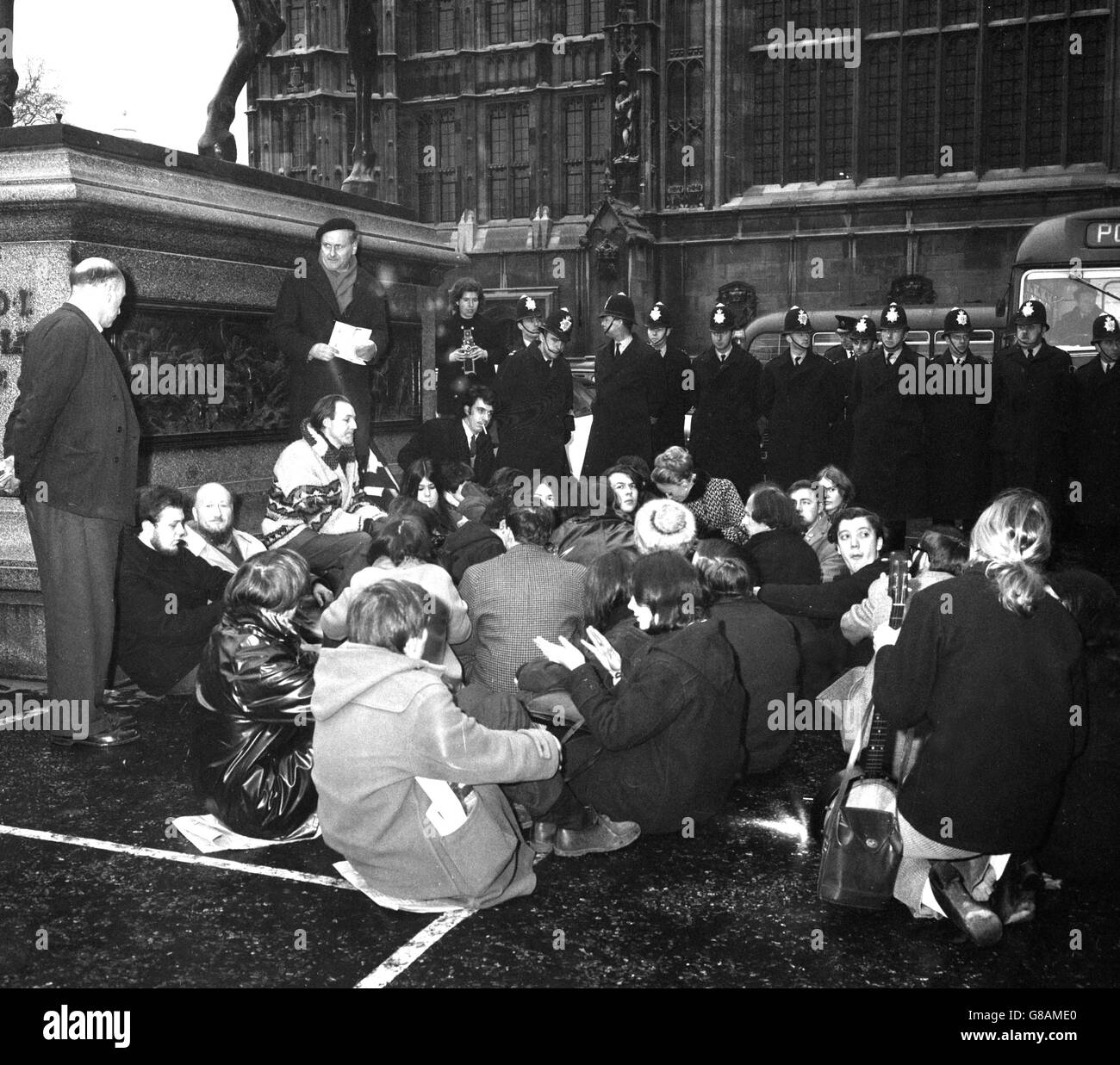 A solid line of police stands above demonstrators seated on the pavement by the Coeur de Lion statue outside the Houses of Parliament in London. This was a demonstration organised by the London Committee of 100 against the war in Vietnam. Though it was intended to hold the meeting in the House of Commons, only three people managed to get in before being escorted away. Police finally ended the sit down by carrying a number of demonstrators into a van. A number of people were charged with obstructing the highway. Stock Photo