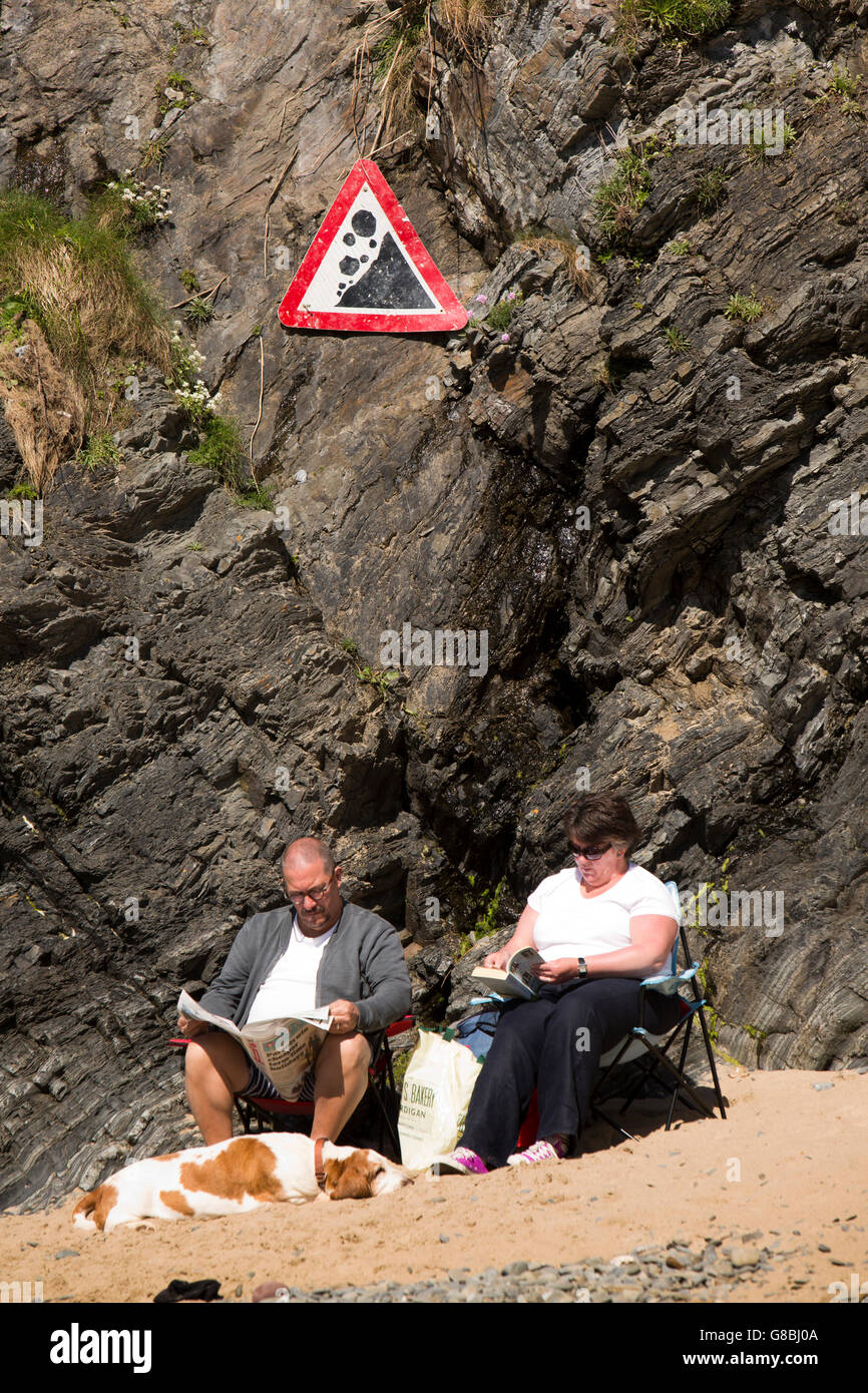 UK, Wales, Ceredigion, Llangrannog, amusing signs, couple sat at base of cliffs ignoring falling rocks danger sign Stock Photo