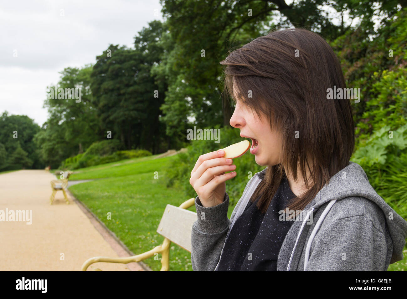 Young  woman, adult or late teens, seated in park eating a slice of apple. Stock Photo