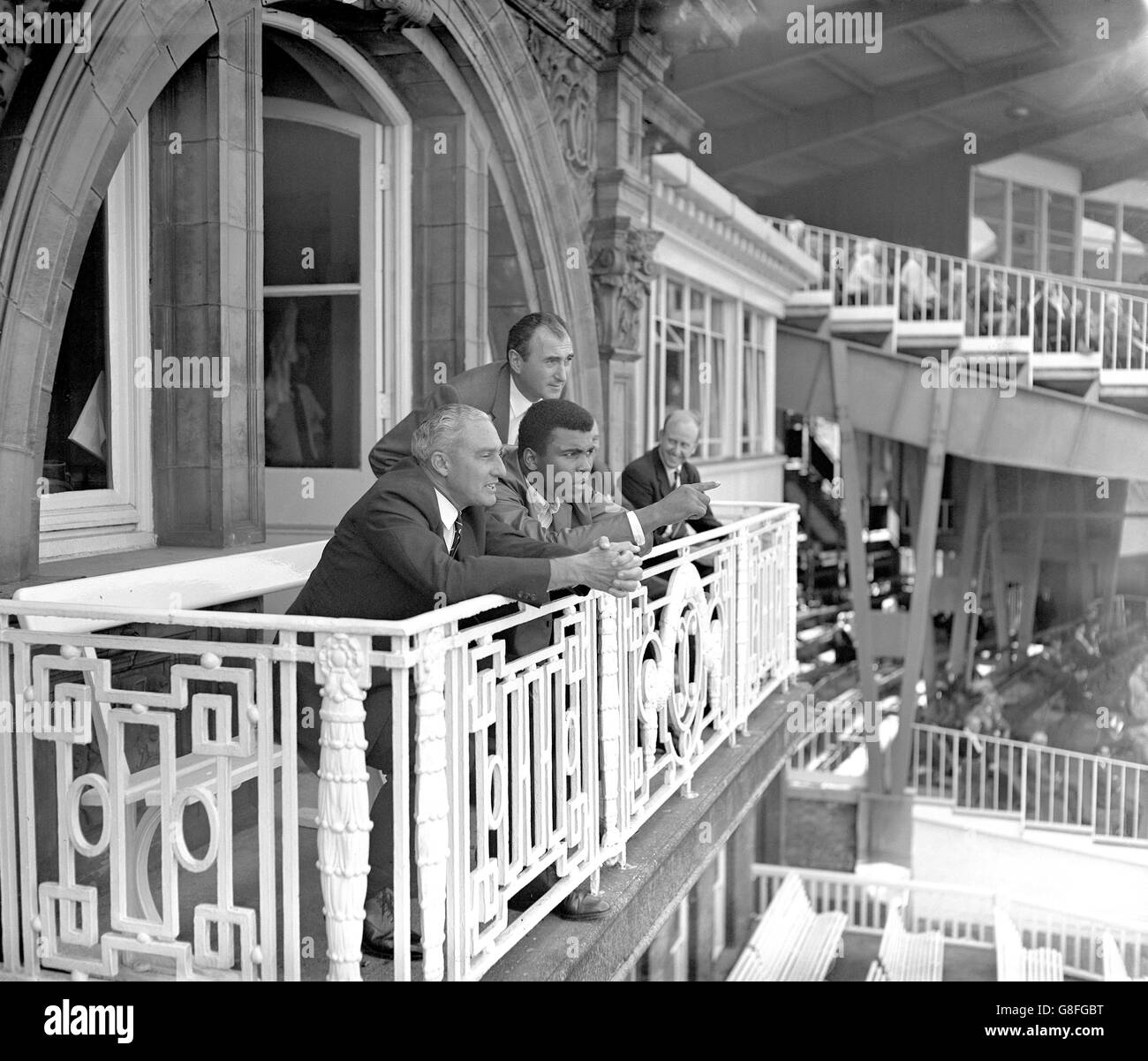 World Heavyweight Boxing Champion Muhammad Ali (second l) quizzes West Indies team manager Jeffrey Stollmeyer (l) about the match while taking in the view from the West Indian dressing room balcony. Helping out with the explanations is Jarvis Astaire (behind Ali), Managing Director of View Sport Ltd Stock Photo