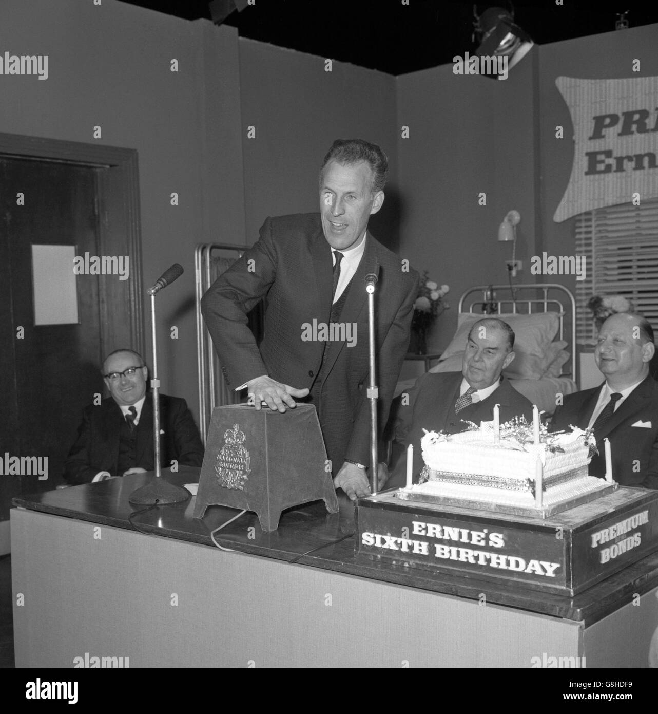 Television entertainer Bruce Forsyth pushes the button to start by remote control 'Ernie', the Premium Bonds electronic selection device based at Lytham St Annes, Lancashire, during a birthday ceremony at the ATV Studios, Elstree, Hertfordshire. Even a special cake had been baked to celebrate the sixth birthday of the Premium Bonds scheme. Looking on (centre) is Viscount Mackintosh of Halifax, chairman of the National Savings Committee. Stock Photo