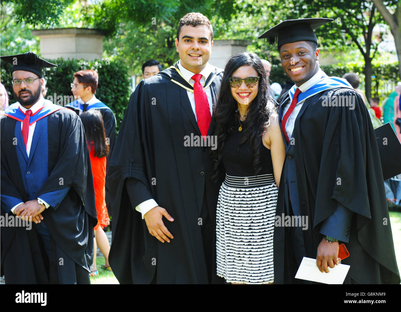 Joyful graduating students in professional gowns posing with lovely girlfriend with long black hair on Graduation Day at Queen Mary London University. Stock Photo