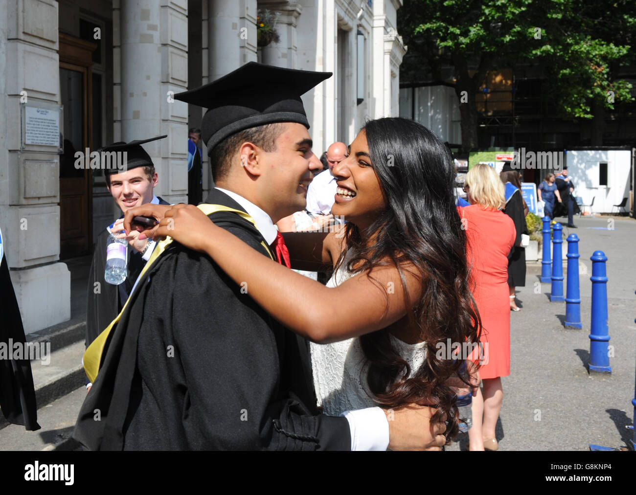 Girlfriend wholeheartedly congratulating and hugging with loving smiles her graduating friend in full gown with hat at Queen Mary London University. Stock Photo