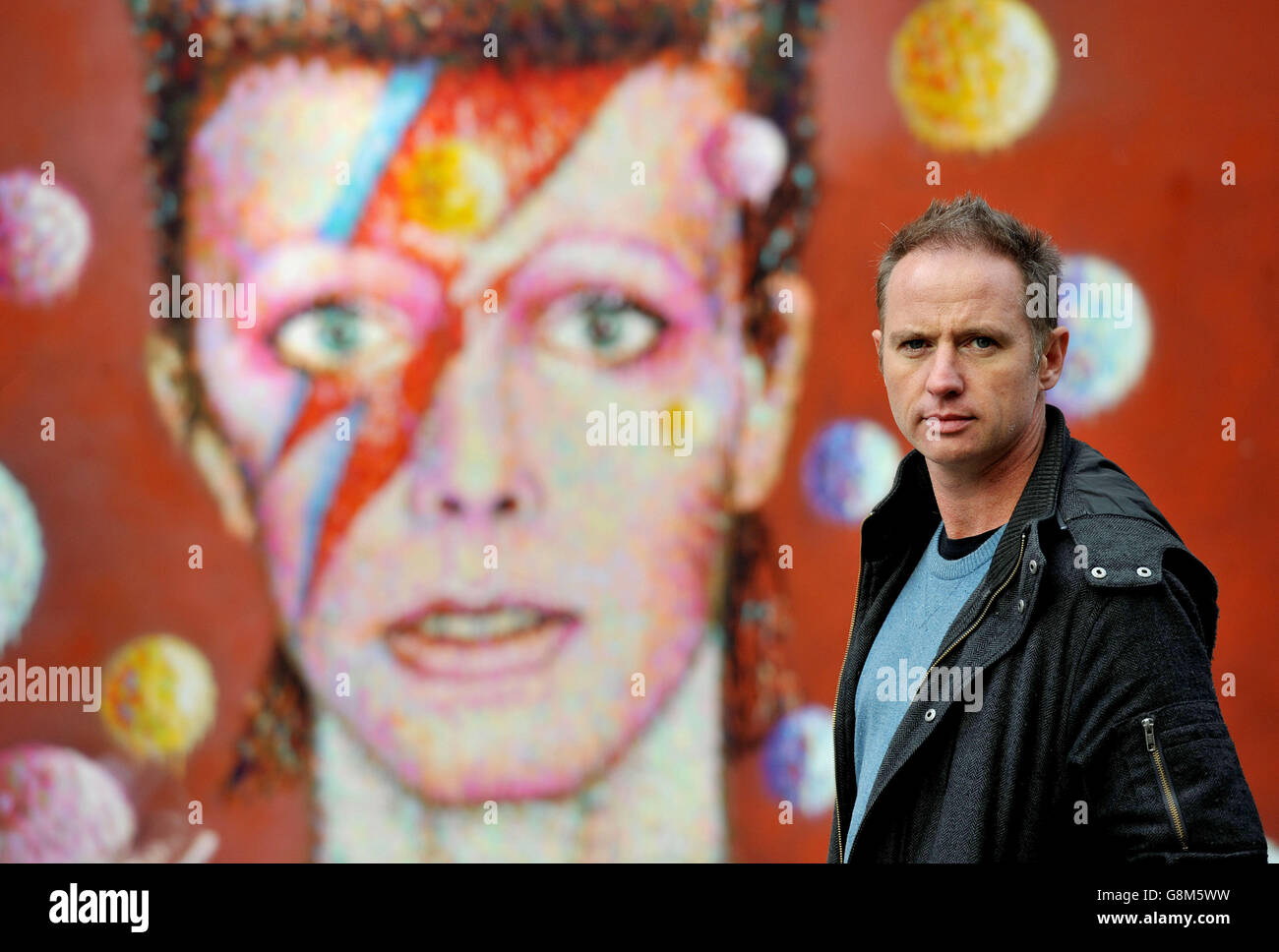 Artist Jimmy C, stands in front of his David Bowie mural artwork on the wall of a Morley's store in Brixton, London, as he views flowers and tributes for the first time after returning from aboard, which have been left for the singer who died of cancer earlier this year. Stock Photo
