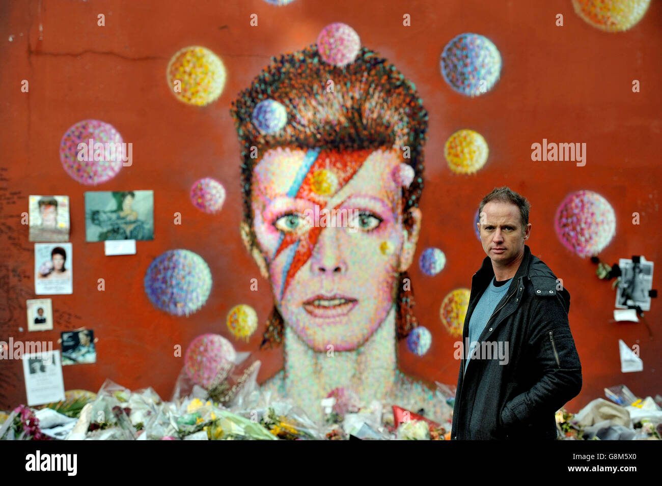 Artist Jimmy C, stands in front of his David Bowie mural artwork on the wall of a Morley's store in Brixton, London, as he views flowers and tributes for the first time after returning from aboard, which have been left for the singer who died of cancer earlier this year. Stock Photo