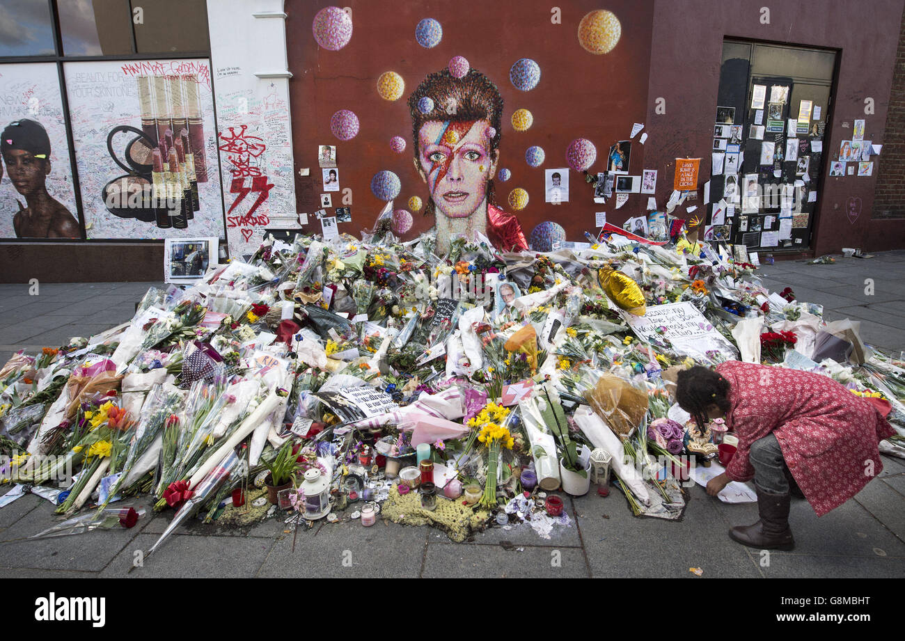 A young girl leaves a tribute in front of a mural of David Bowie on the wall of a Morley's store in London's Brixton, the singer's birthplace, almost three weeks after his death. The star wanted his ashes to be scattered in a Buddhist ritual in Bali, according to his will. Stock Photo