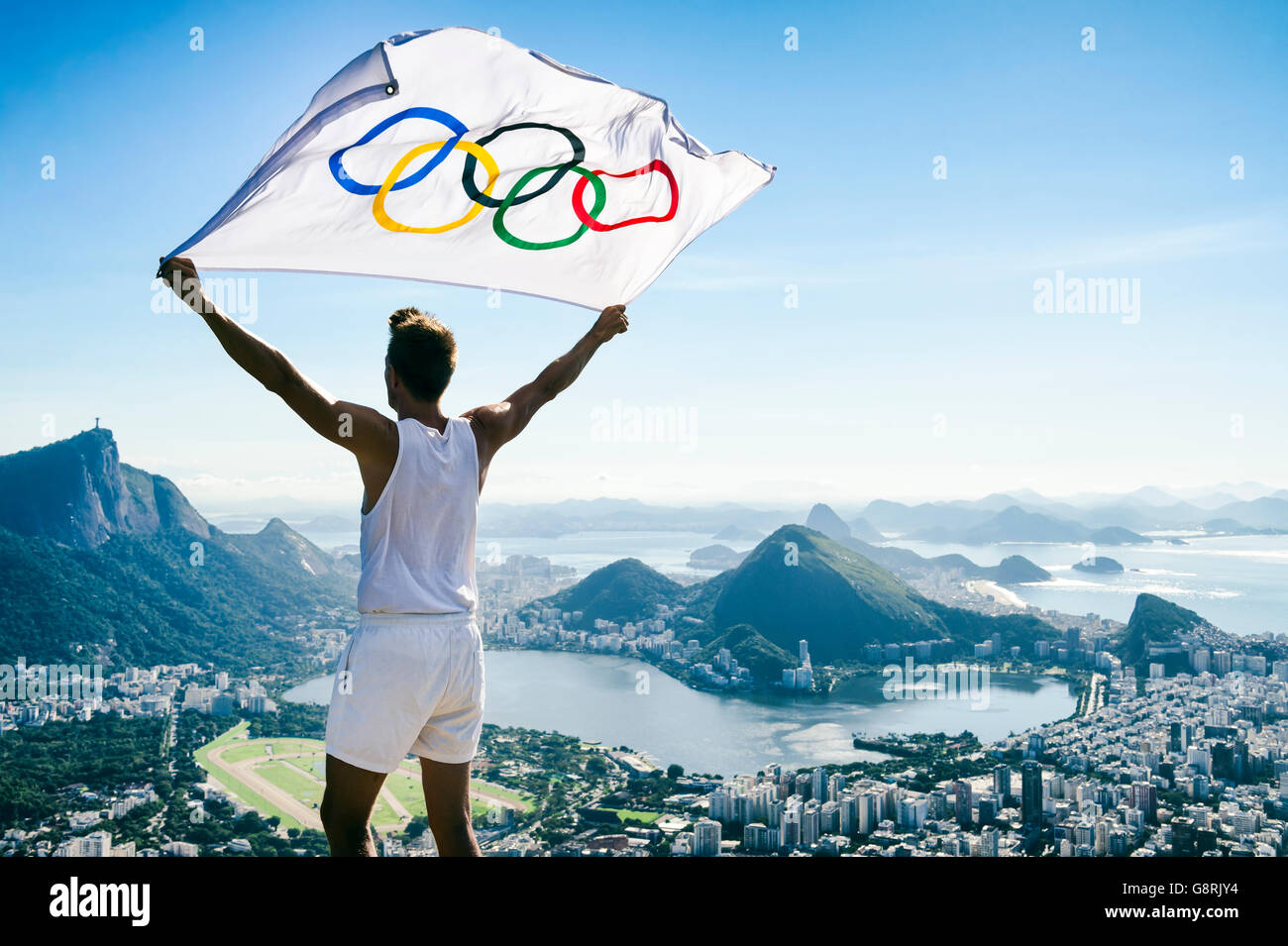 RIO DE JANEIRO - MARCH 21, 2016: Athlete stands holding Olympic flag above a city skyline view of Corcovado Mountain. Stock Photo