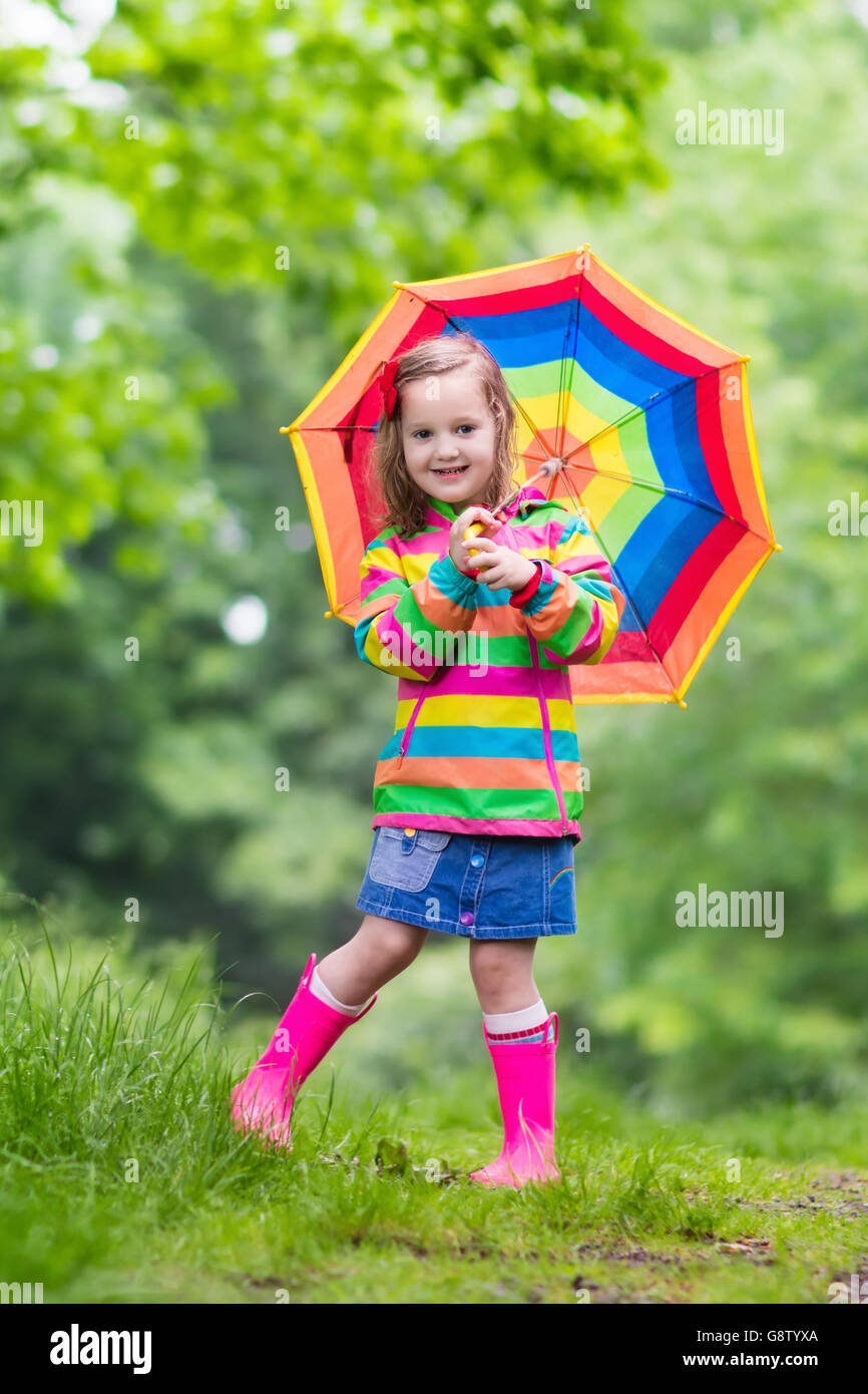Little girl playing in rainy summer park. Child with rainbow umbrella waterproof coat and boots jumping in puddle in the rain Stock Photo Alamy