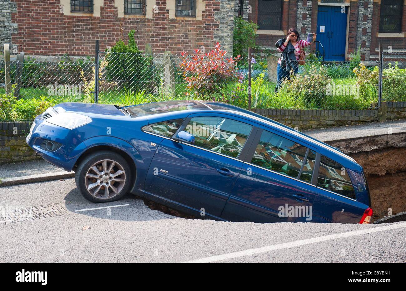 A woman takes a photograph of a car which has partially disappeared down a sinkhole in Woodland Terrace in Greenwich, south-east London. Stock Photo
