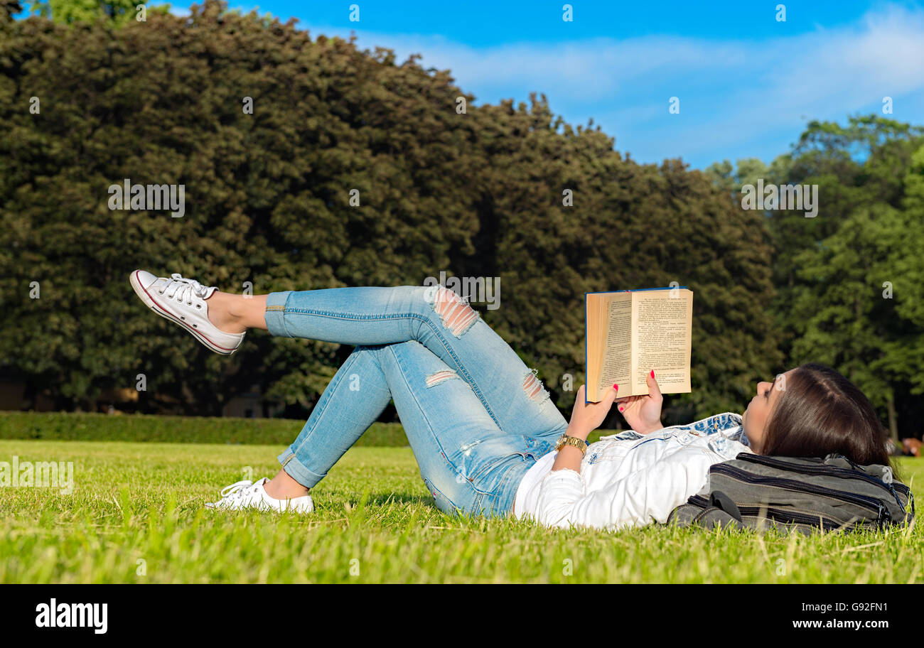 Smiling beautiful young woman on grass and reading book, against background of summer green park. Stock Photo