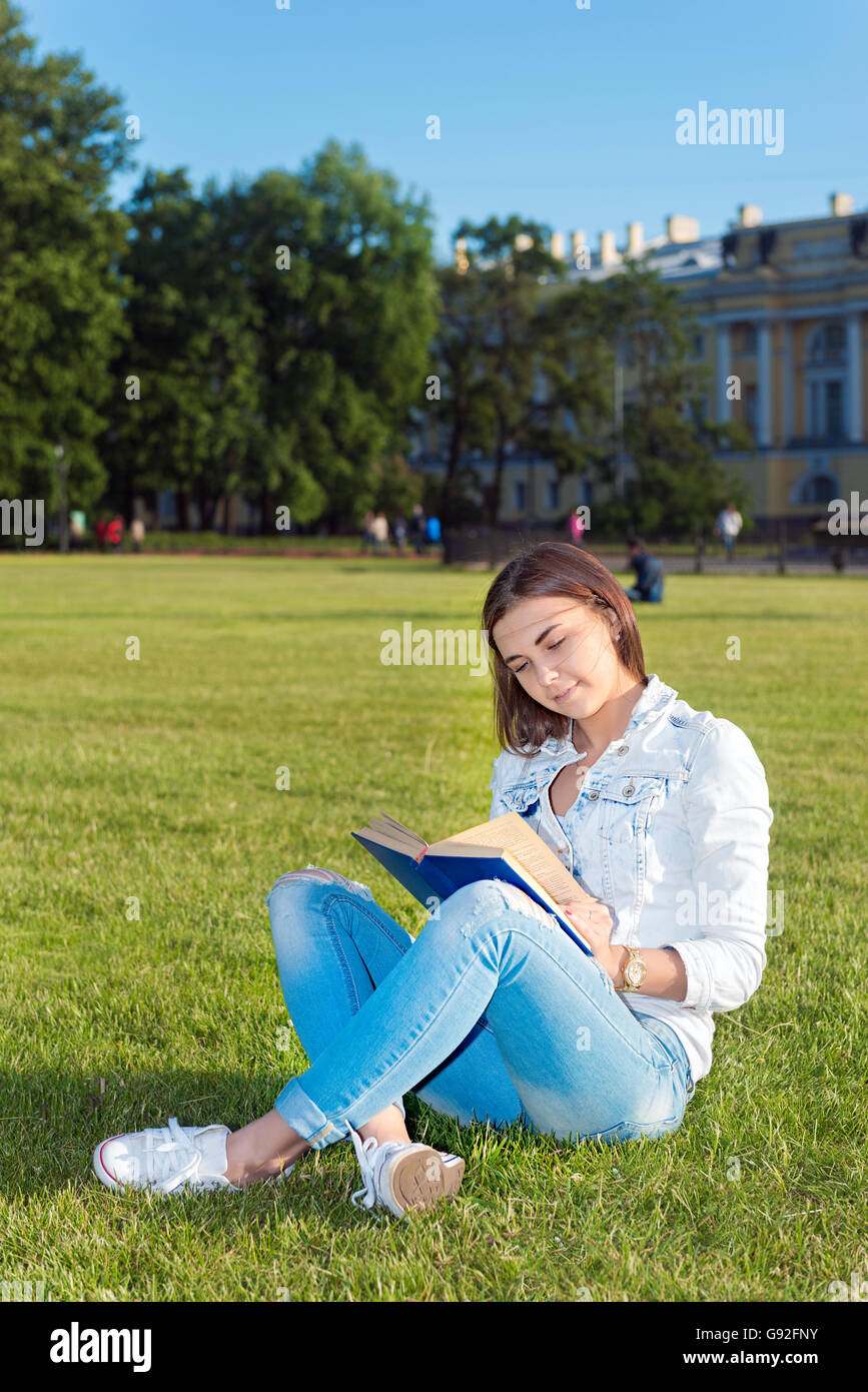 Smiling beautiful young woman on grass and reading book, against background of summer green park. Stock Photo