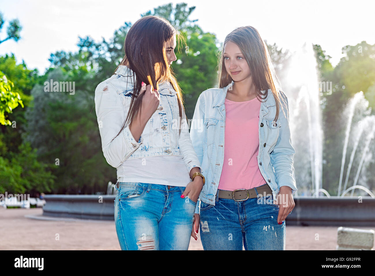 Two beautiful girls walking in the summer park end talking. Friends wearing stylish shirt and jeans , enjoying day off and have Stock Photo