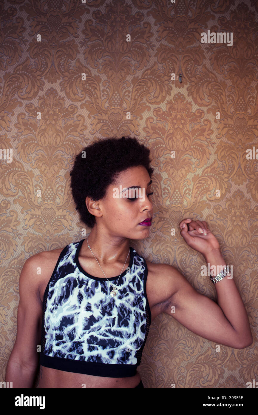 Beauty in dereliction : Fashion photography of a young afro-caribbean woman girl alone in an ornately  wall-papered room  wearing fitness-style clothes, looking sultry and moody Stock Photo