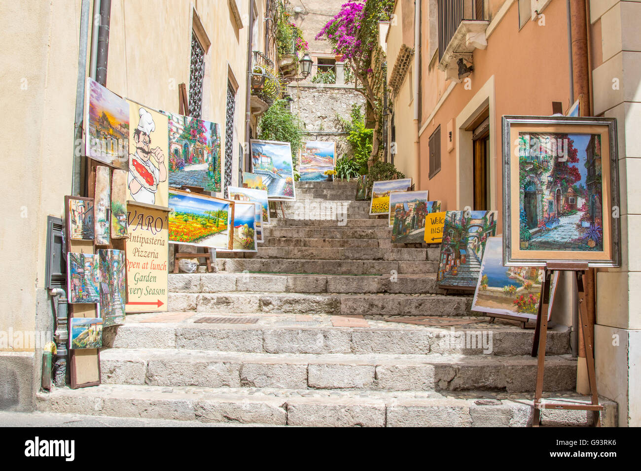 Street scene in Taormina, Italy. Paintings for sale displayed either side of cobbled staircase leading off Corso Umberto Stock Photo