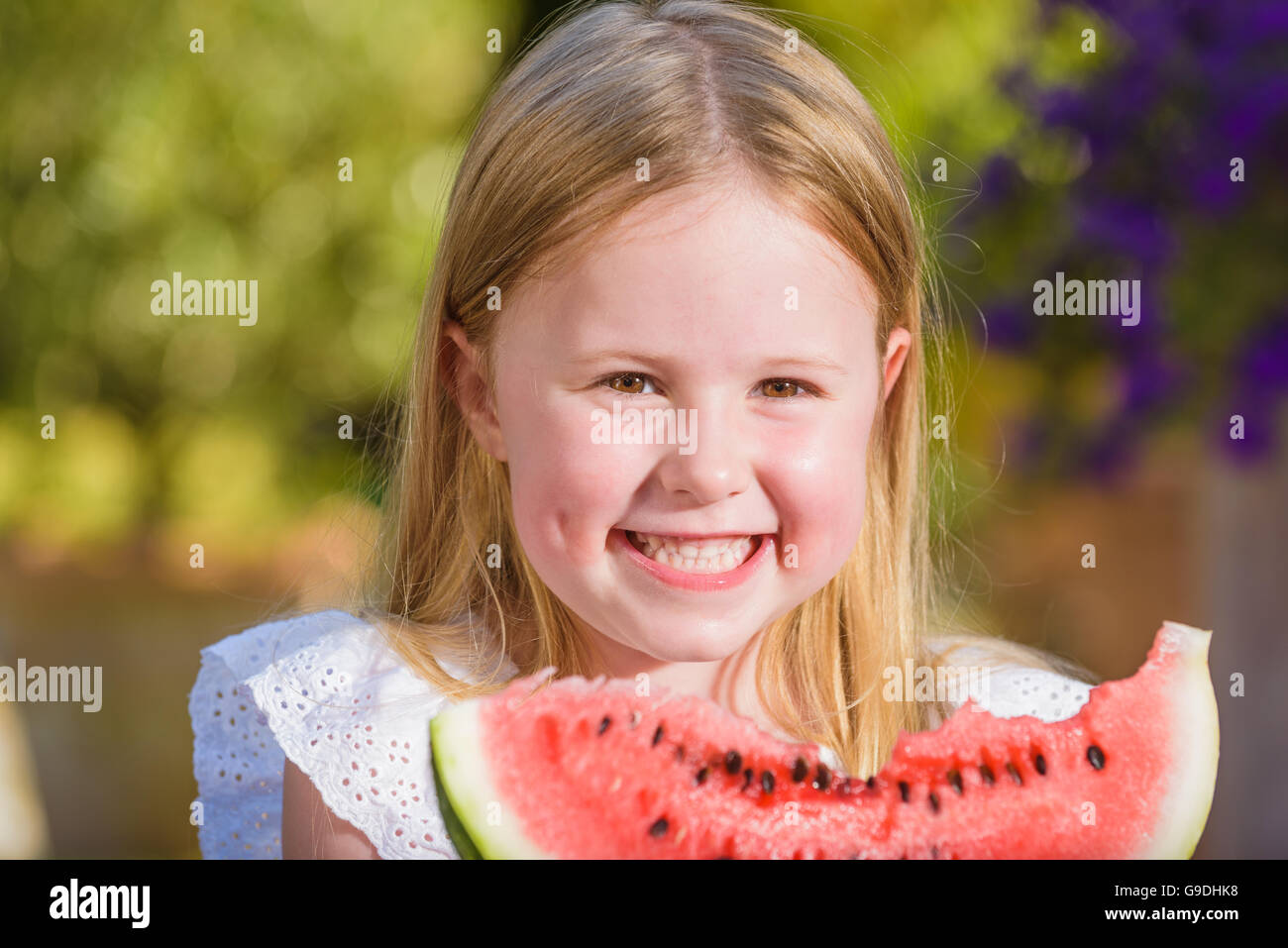 Happy girl with big red slice of watermelon sitting on rustic tabels in summer garden. Stock Photo