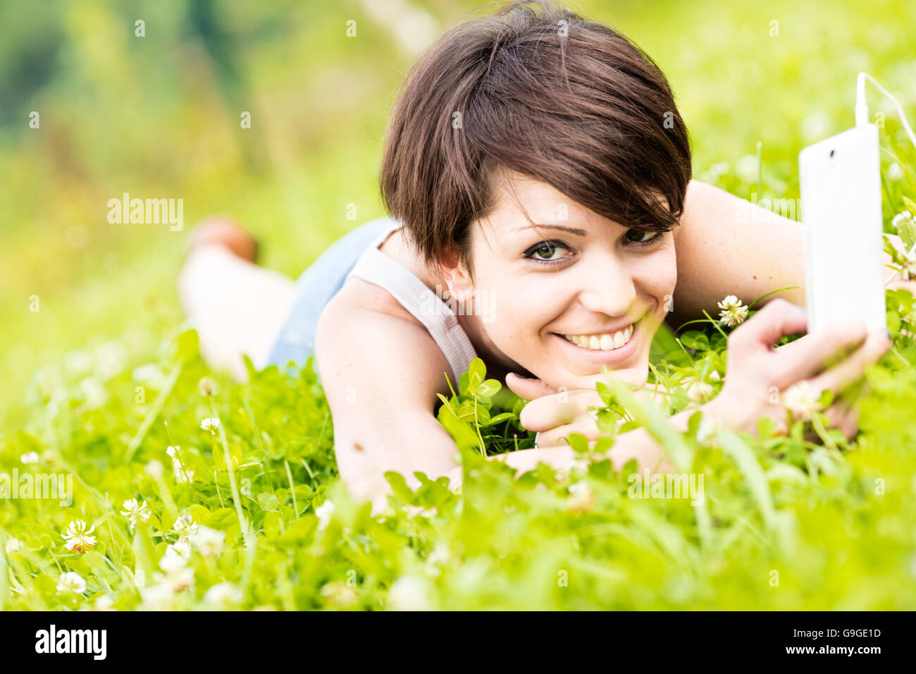 Young woman lying on her stomach in fresh spring grass listening to music on her mobile phone smiling at the camera with pleasur Stock Photo