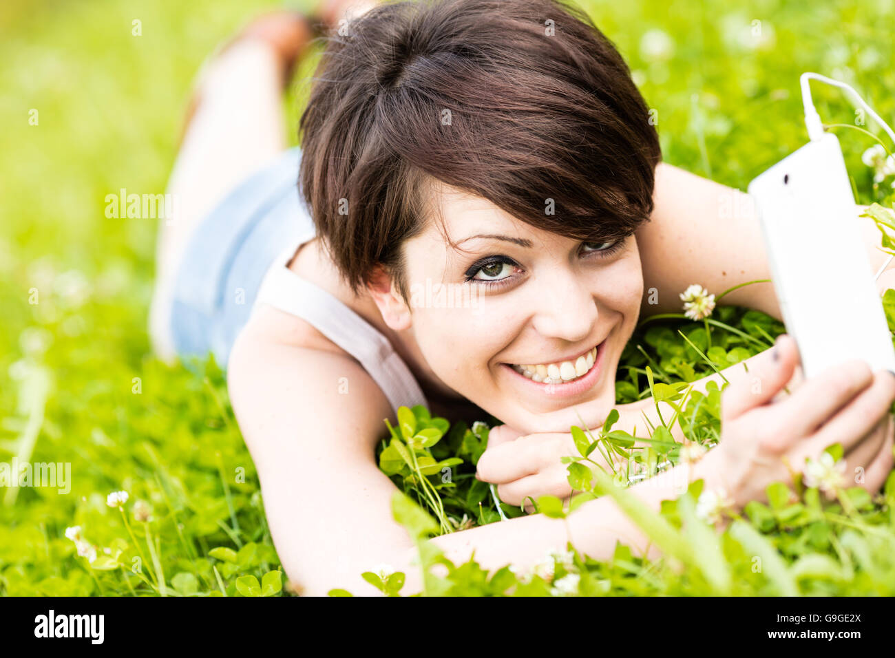 Pretty happy woman relaxing on her stomach in fresh spring grass in a meadow with her mobile phone listening to music with a bli Stock Photo