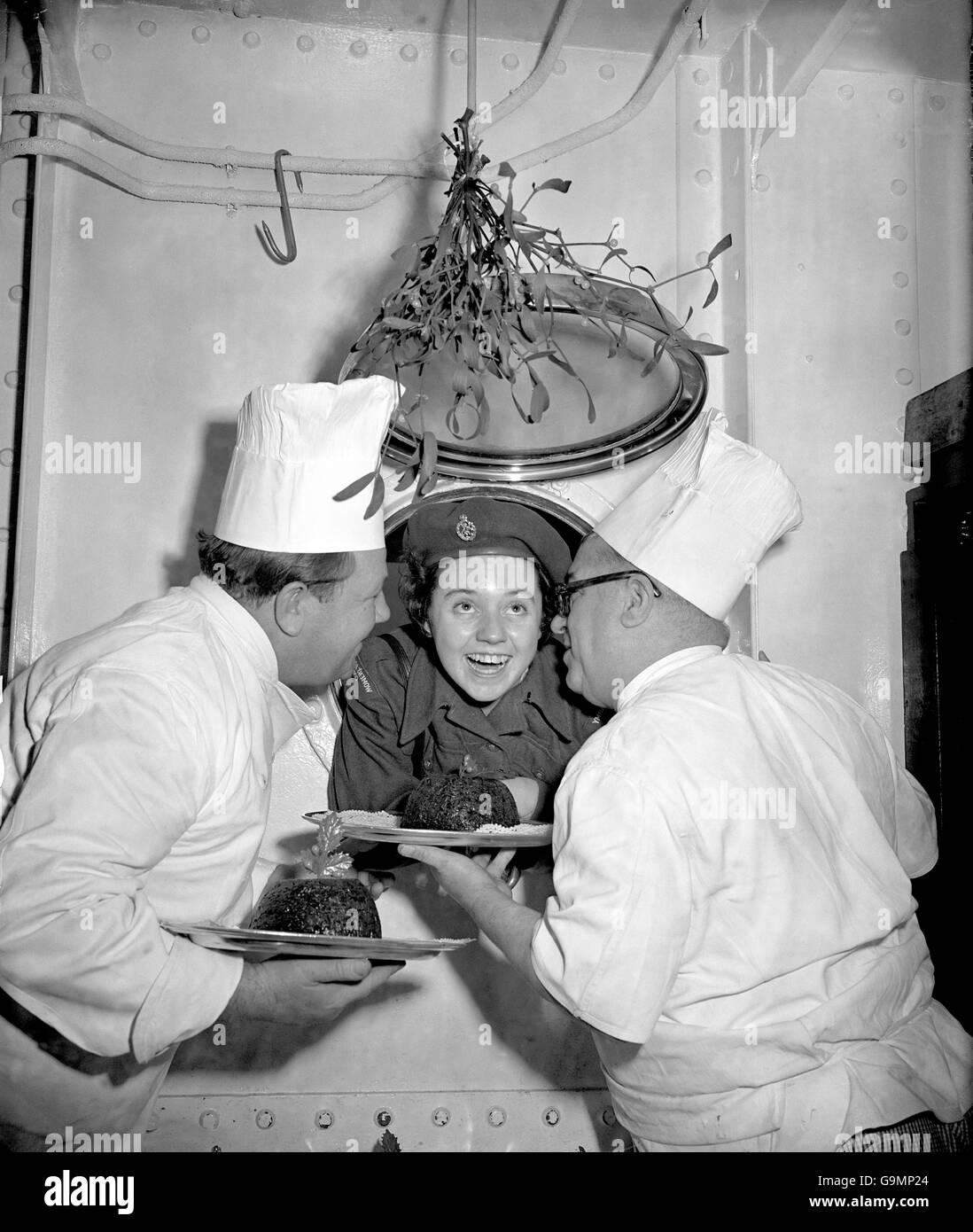 Making herself a fine target for a kiss beneath the mistletoe is WRAC Private Anne Hughes, who is on the way to Singapore aboard the troopship HMT Dunera, which is currently stationed at Southampton. She is peeping through the porthole as Chief baker Charles Taylor (right) and his assistant Henry Mallett prepare Christmas puddings. Stock Photo