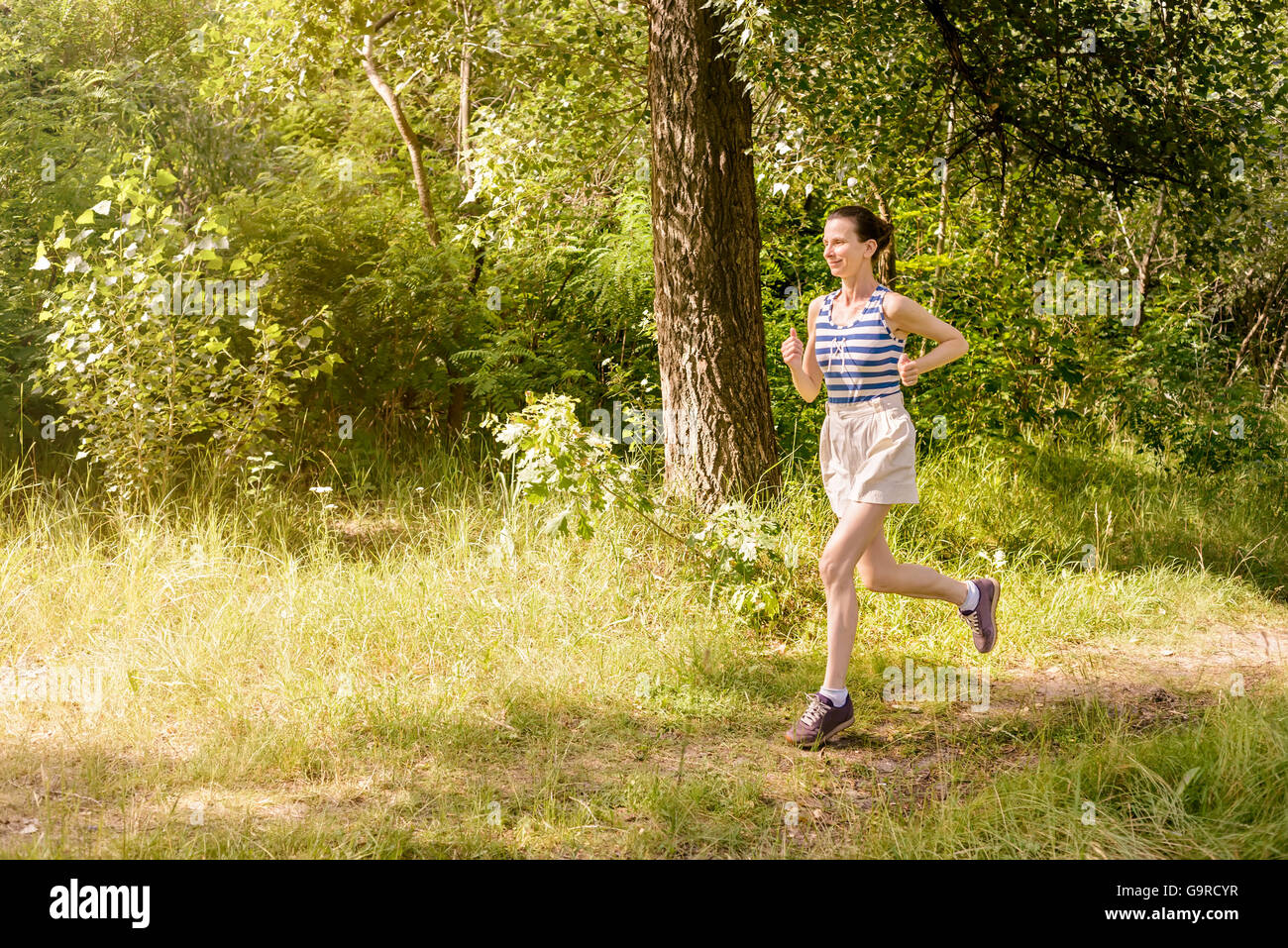 A happy senior woman is running toward the light in the forest during a warm summer day Stock Photo