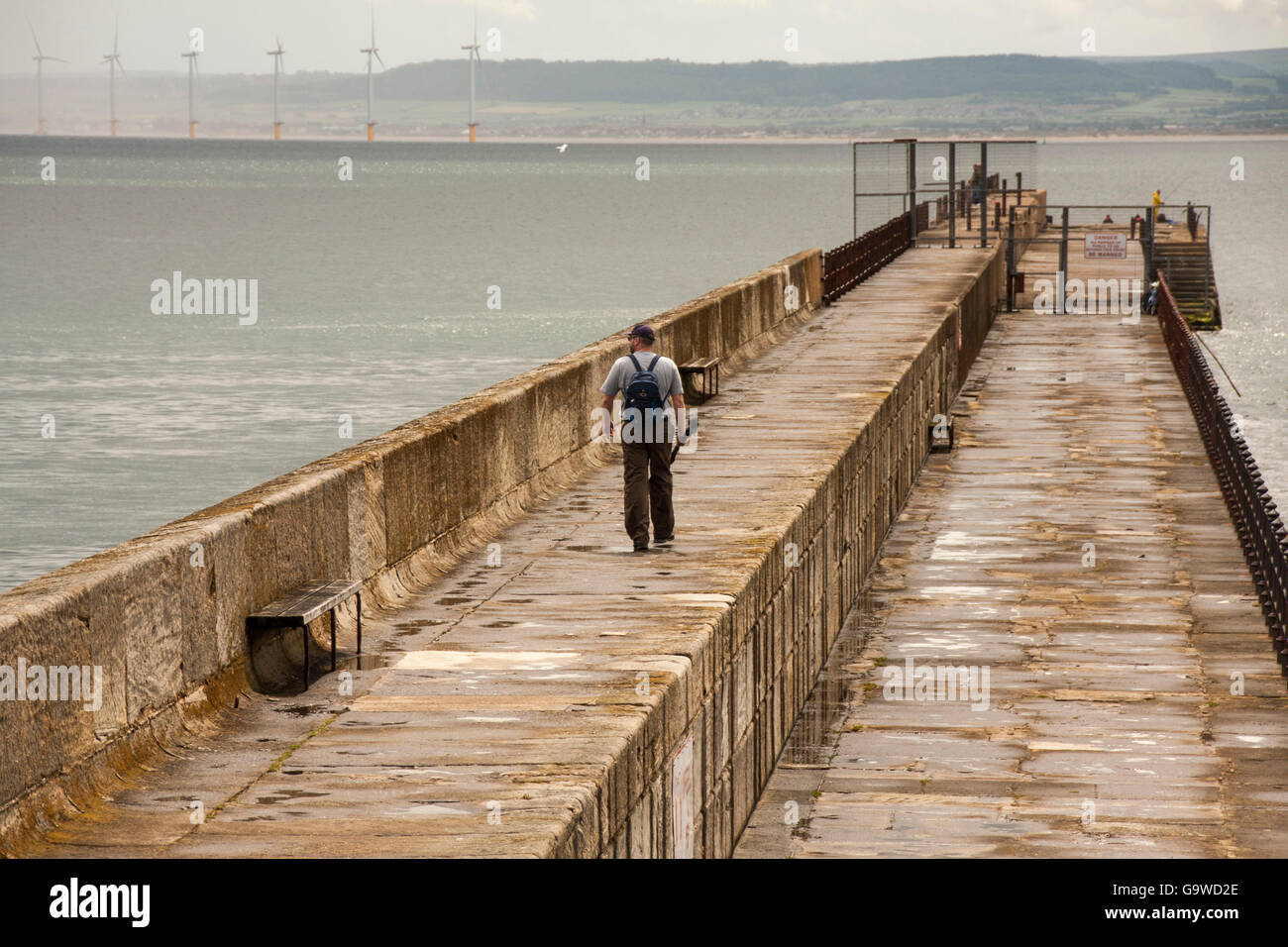 A man walking along the Headland at Hartlepool carrying his rod  for a days fishing with the wind turbines in the background Stock Photo