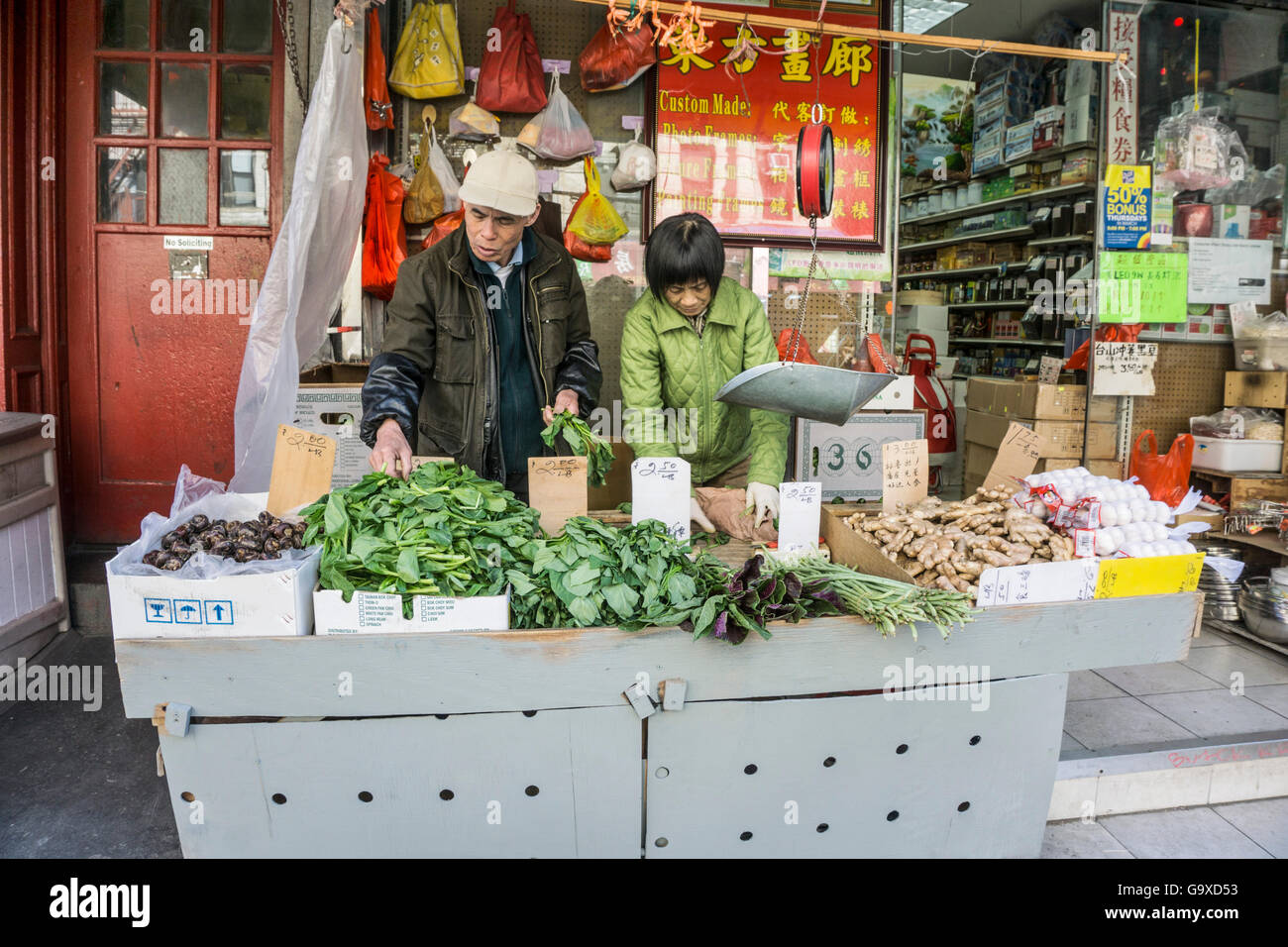 elderly Chinese couple tending their vegetable stall on Mott street sidewalk outside colorful mom & pop general store Chinatown Stock Photo