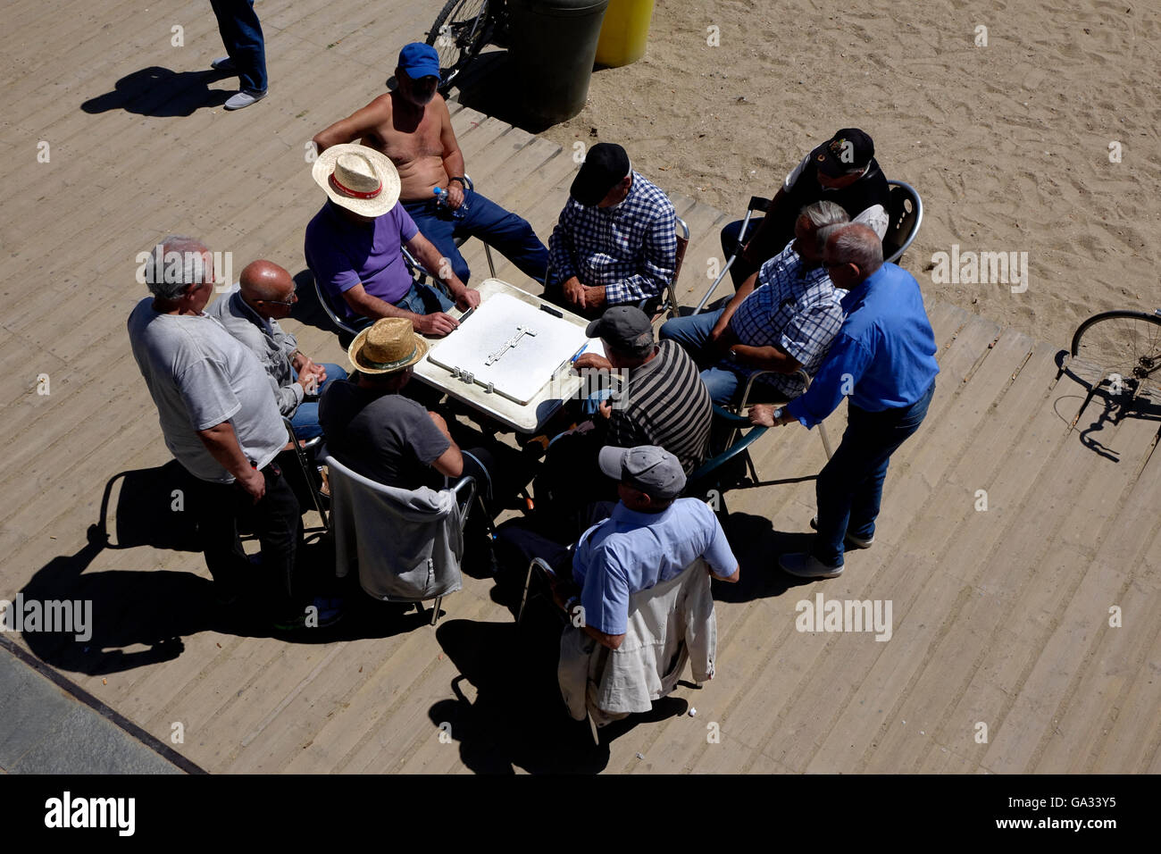 A group of men enjoy watching a game of dominos on the boardwalk at Barcelona's Platja Nova Icaria. Stock Photo
