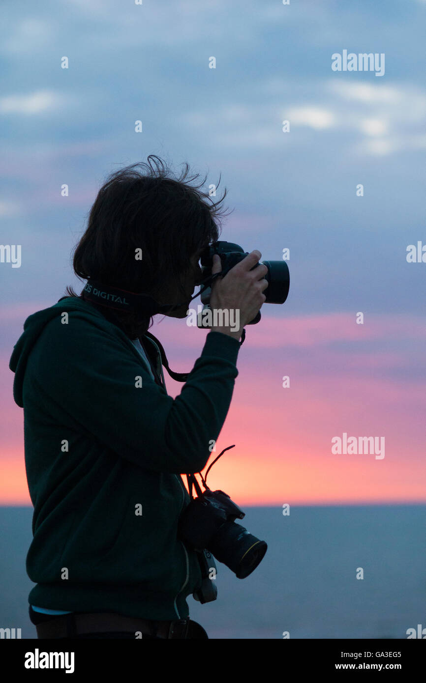 A silhouette of a professional photographer man taking a photograph with a DSLR camera against a sunset Stock Photo