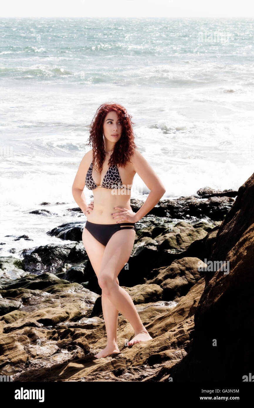 Hispanic Woman Outdoors On Rocks In Bikini Full-Length With Ocean In Background Arms Akimbo Stock Photo