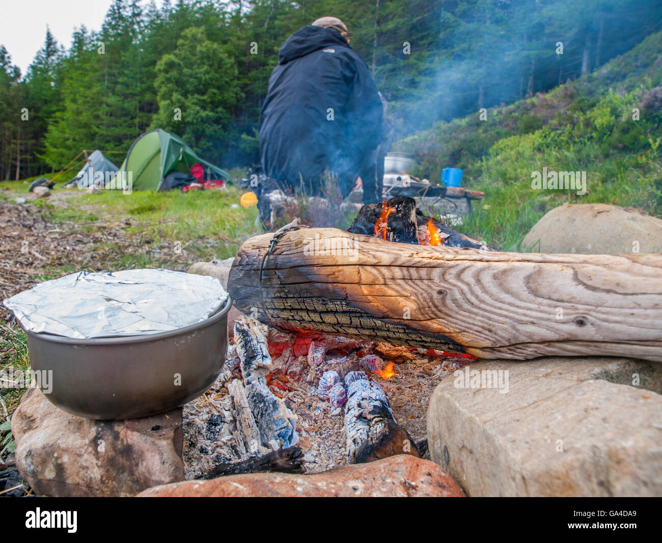 A cooking pan on a camp fire, camper and tents in background. Scottish