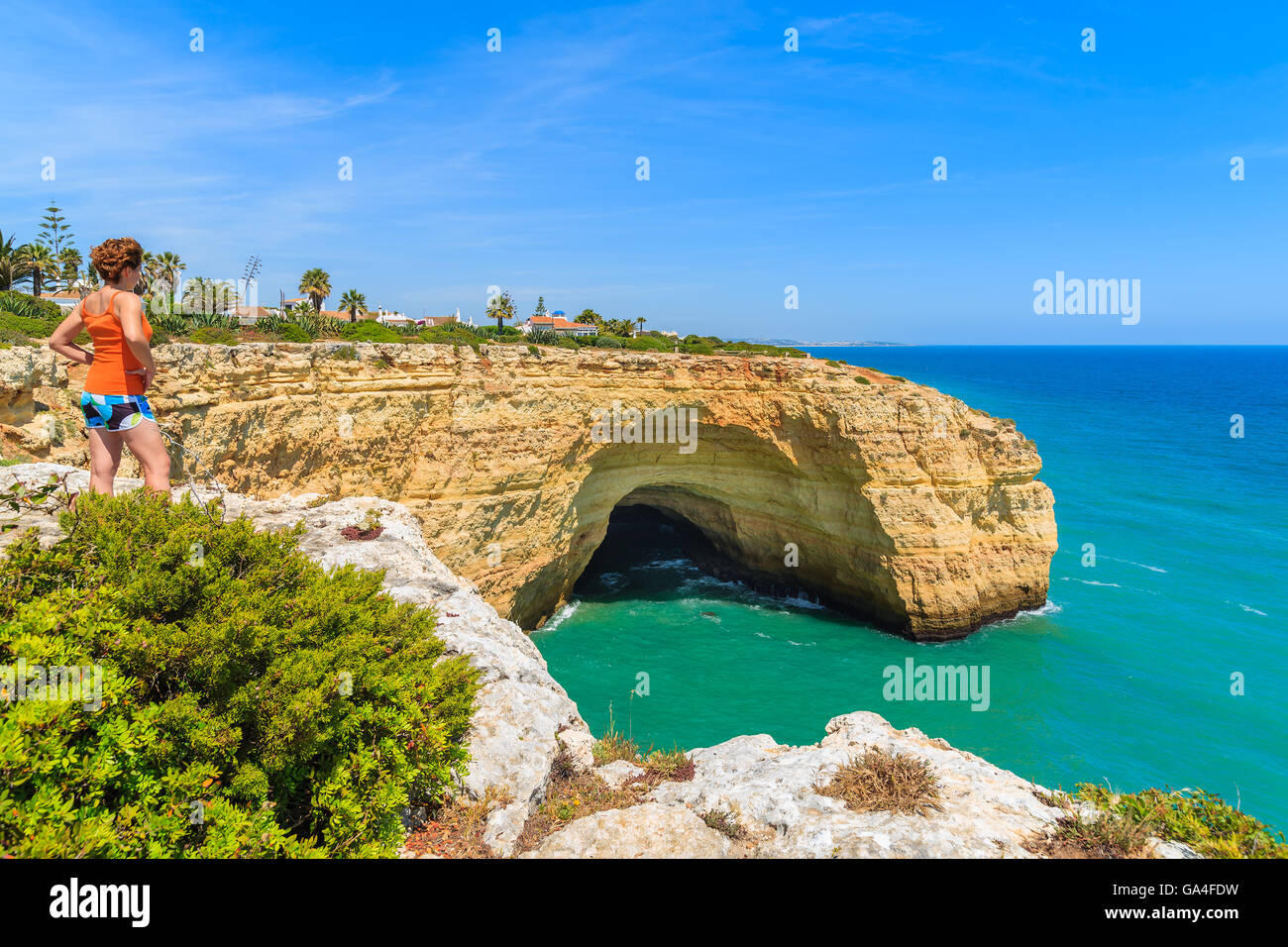 Young woman tourist standing on cliff rock and looking at sea cave on coast of Portugal, Algarve region Stock Photo