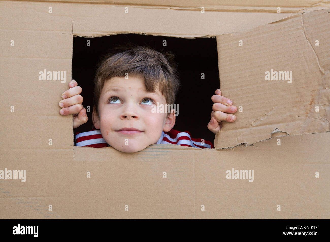 A 7 year old boy looks out of a window cut from a cardboard box Stock Photo