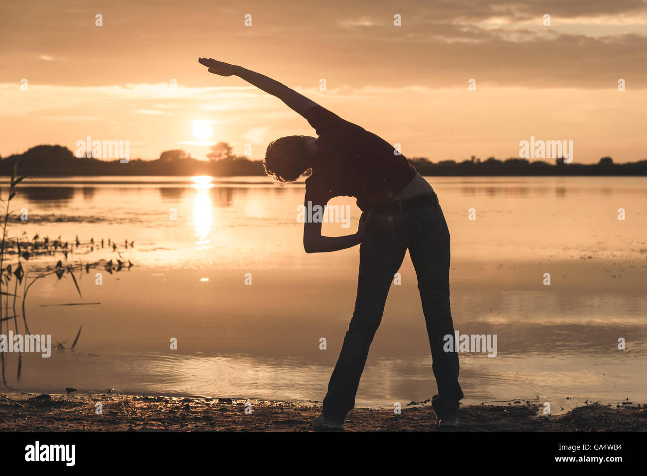 young slim woman doing exercises on the shore of lake at sunset Stock Photo