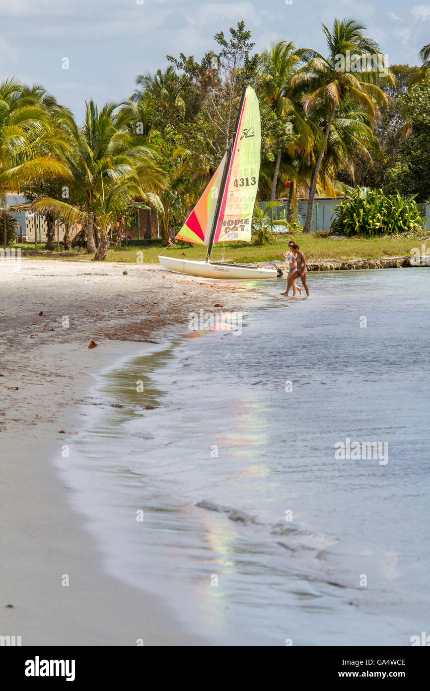 Women in bikinis walking along beach, with Hobie Cat in background, at Playa Larga, Matanzas, Cuba Stock Photo