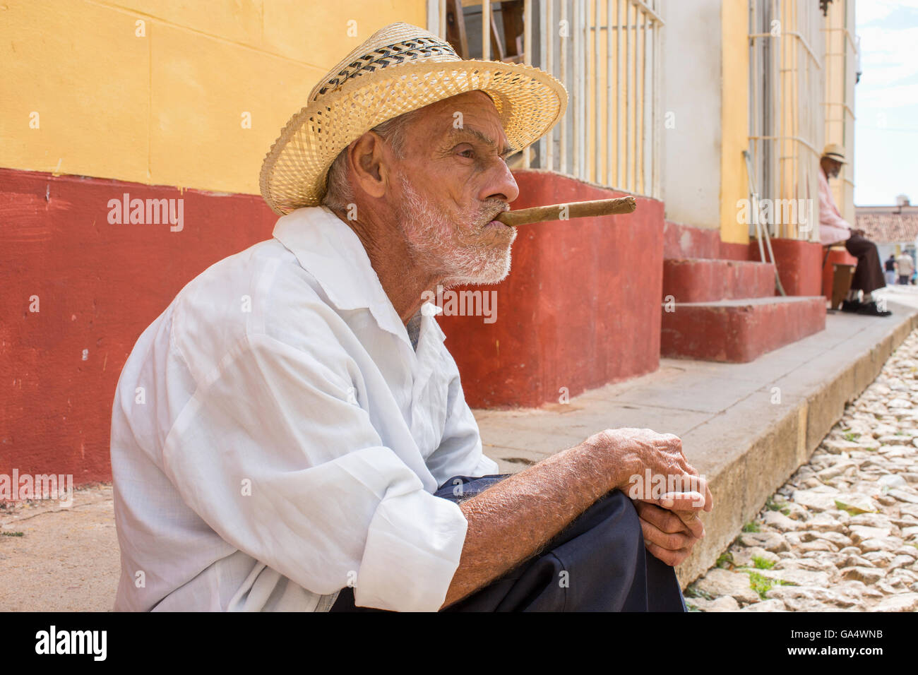 Local man in hat with cigar sitting on sidewalk in Plaza Major, Trinidad, Cuba Stock Photo