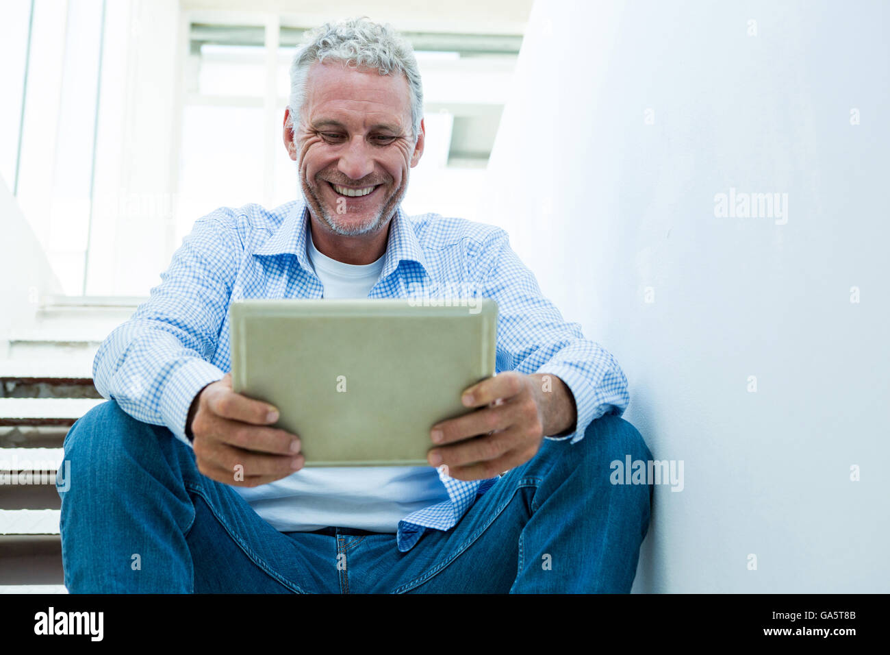 Smiling mature man holding tablet while sitting on steps Stock Photo