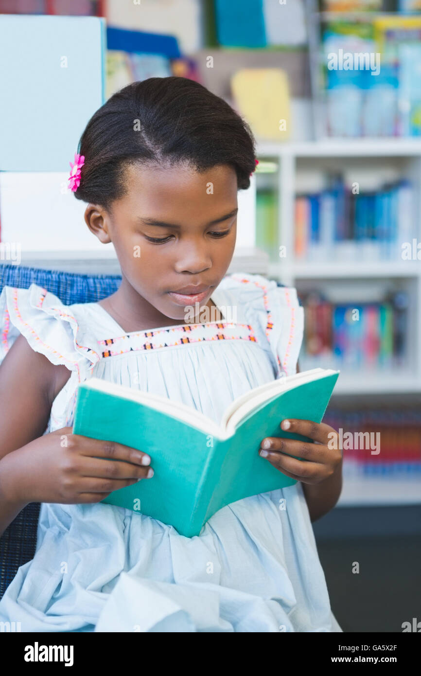 Schoolgirl sitting on chair and reading book in library Stock Photo