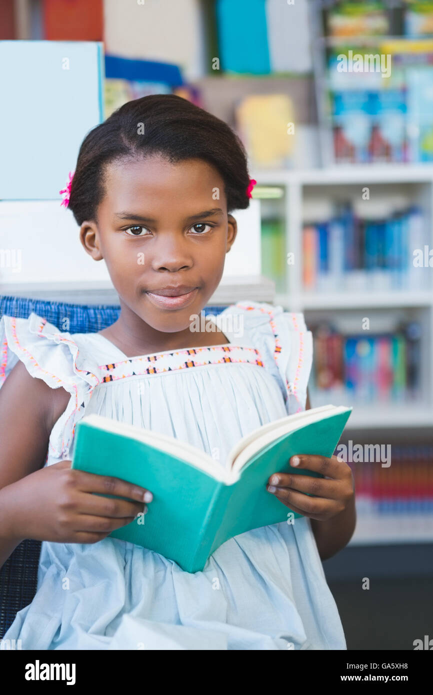 Schoolgirl sitting on chair and reading book in library Stock Photo