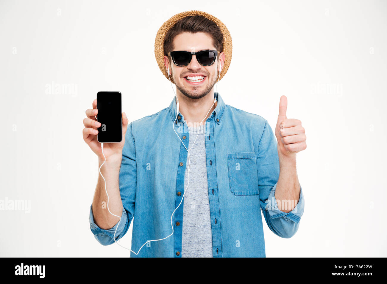 Cheerful young man in earphones showing blank screen cell phone and showing thumbs up over white background Stock Photo