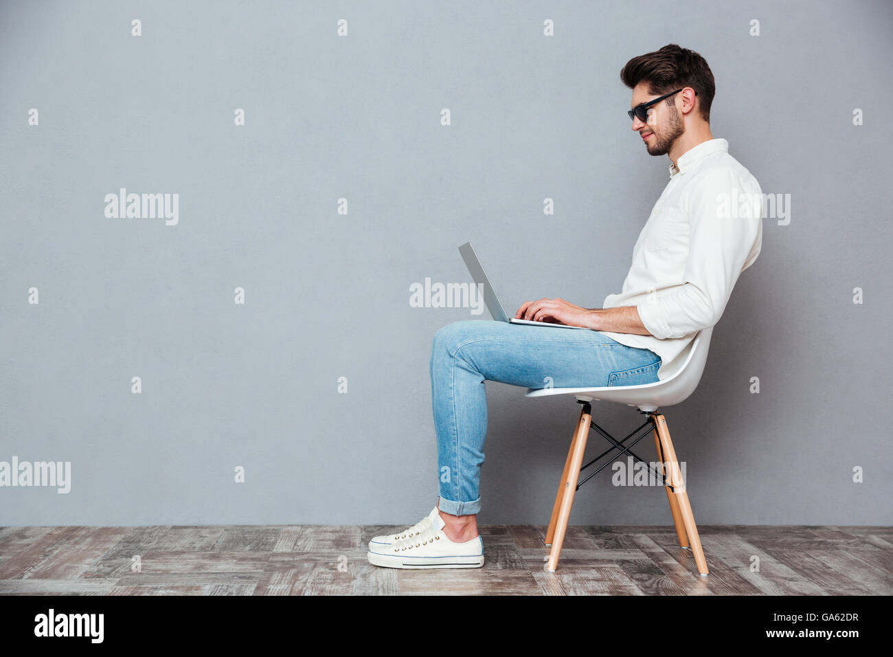 Profile of serious young man in sunglasses sitting on chair and using laptop over grey background Stock Photo