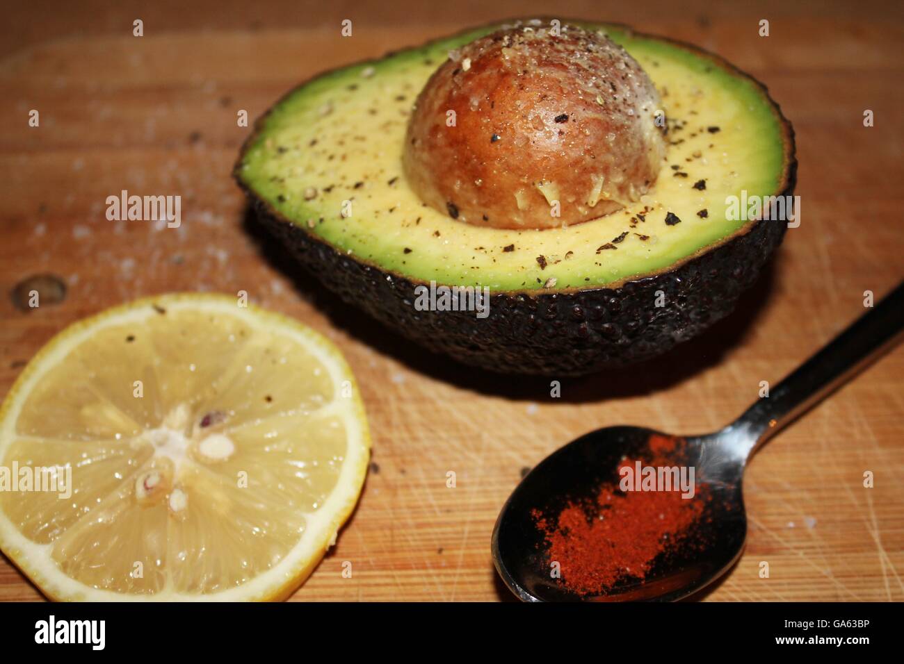 Avocado, stone in, sprinkled with salt and pepper, slice of lemon and spoonful of cayenne pepper at side on wooden board. Stock Photo