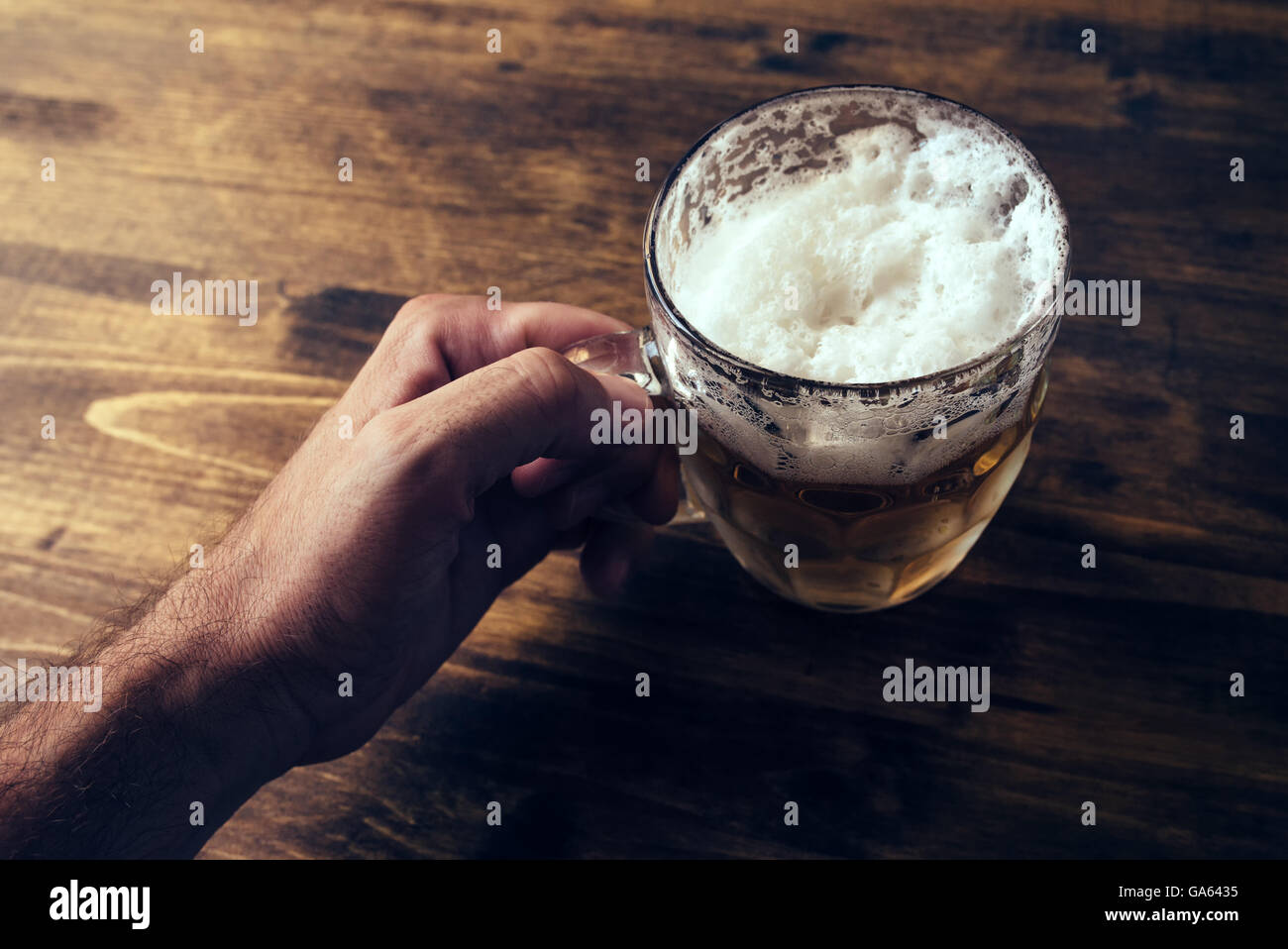 Hand holding beer mug full of cold fresh alcohol drink on wooden background, man in the bar, pov shot, selective focus Stock Photo