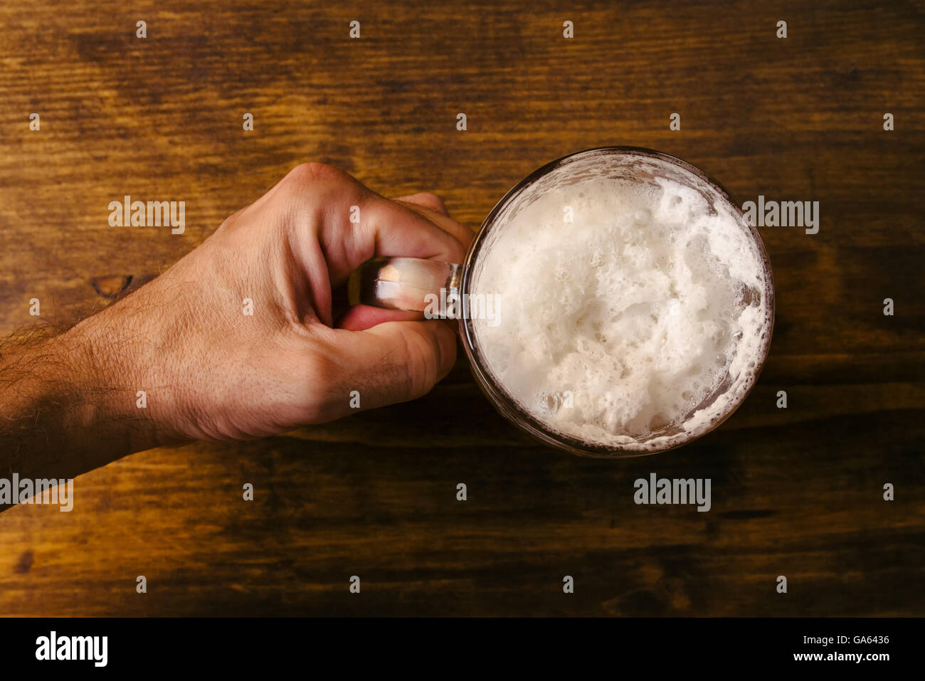 Hand holding beer mug full of cold fresh alcohol drink on wooden background, man in the bar, pov shot, top view, selective focus Stock Photo