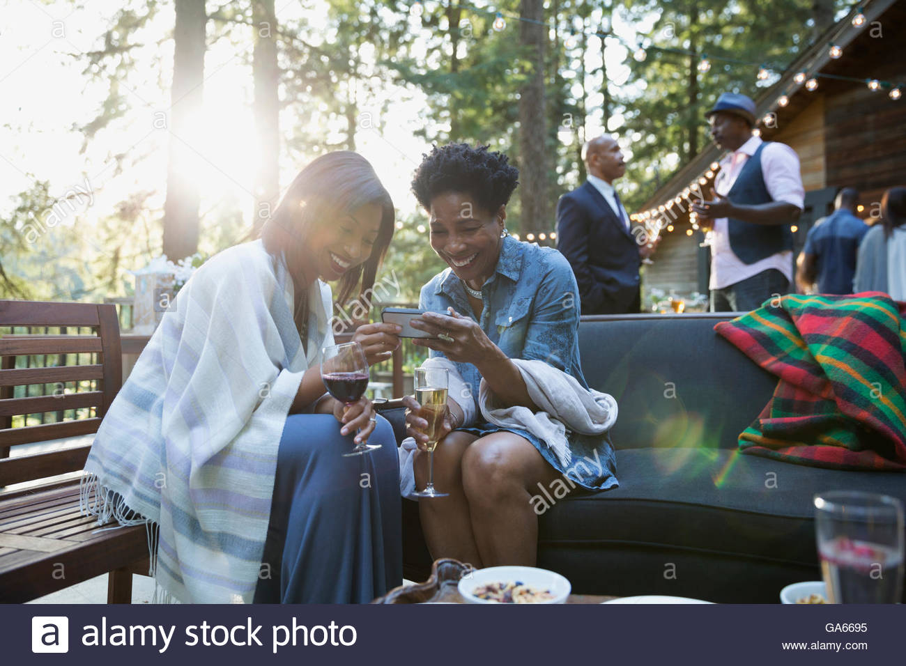Smiling women drinking wine and looking at cell phone at party on balcony in sunny woods Stock Photo