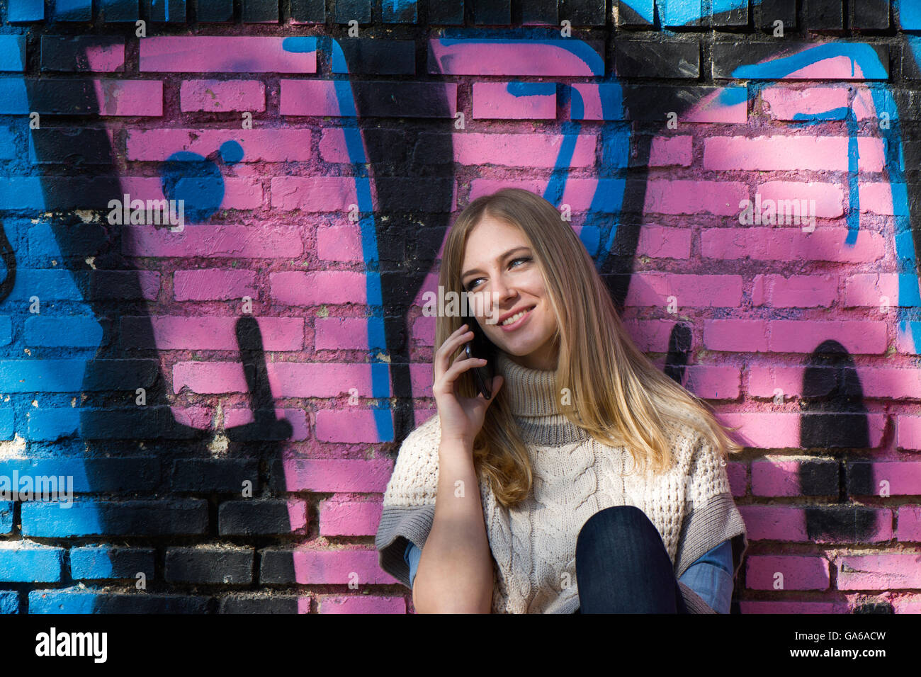 Young blonde woman talking over phone and smiling while sitting on ground against of brick wall with graffiti Stock Photo