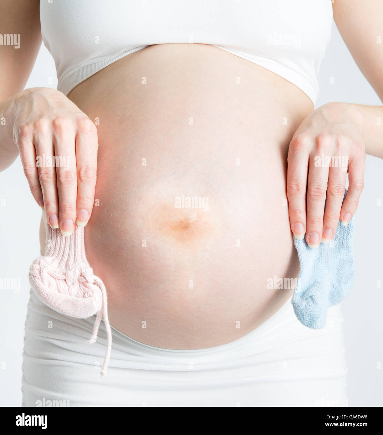 A bregnant woman holds one pink bootie and one blue bootie in front of her baby bump. Stock Photo