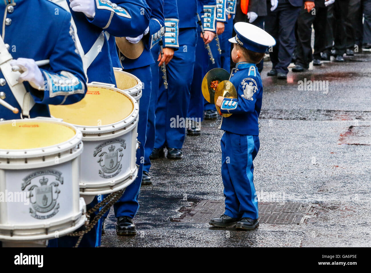 Small boy, in uniform and holding a small set of cymbals, watching the parade of adults walk by, Glasgow, Scotland, UK Stock Photo